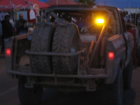 1990 Ford Bronco - Rear of an off-road race truck with dual spare tires and a glowing amber roof beacon at dusk near vendor tents.