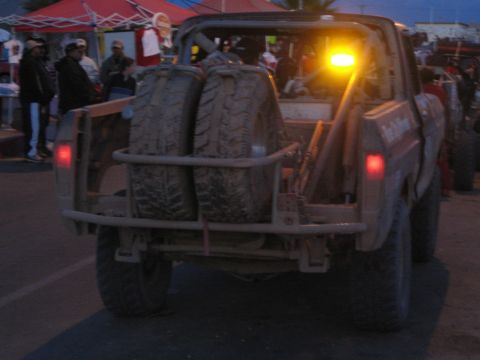 1990 Ford Bronco - Rear of an off-road race truck with dual spare tires and lit amber beacon, surrounded by a crowd at dusk.