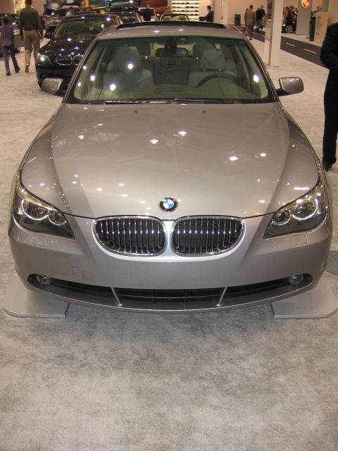 1999 Ford Crown Victoria - A silver BMW sedan is displayed at an auto show, with other vehicles and people in the background.