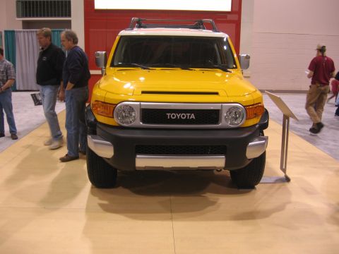 1999 Ford Crown Victoria - A yellow Toyota FJ Cruiser is displayed indoors, with people standing nearby and a sign to its right.