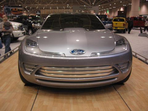 1999 Ford Crown Victoria - A silver concept car is displayed at an auto show with other vehicles in the background.