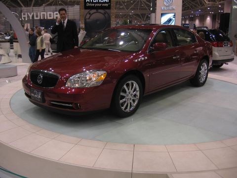 1999 Ford Crown Victoria - A maroon Buick sedan is displayed indoors, with a man in a suit standing behind it and another vehicle in the background.