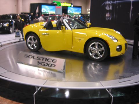 1999 Ford Crown Victoria - Yellow Pontiac Solstice GXP convertible displayed at an auto show with "SOLSTICE GXP" signage.