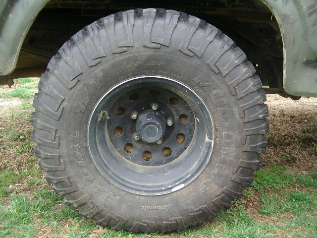 Off-road tire on a black rim, showing part of a green vehicle body and suspension. Text on tire reads "MUD-TERRAIN T/A"