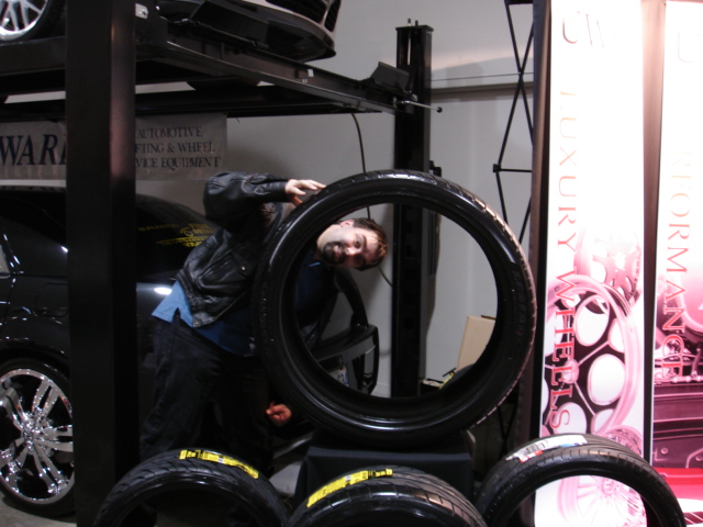 Guy peeking through a tire at a car show, with other tires and displays around