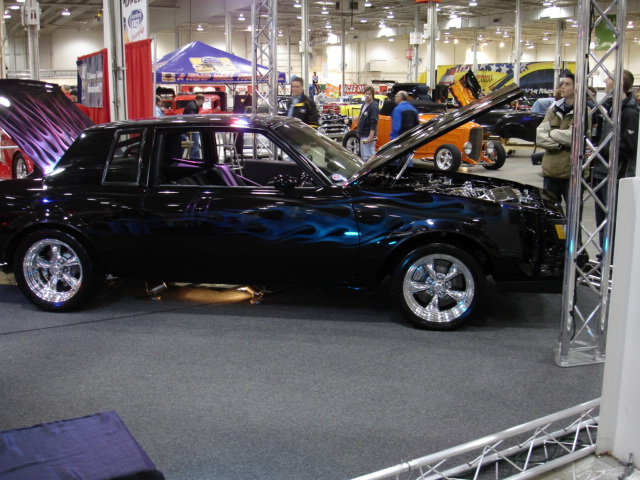 Black coupe with blue flame graphics, hood and trunk open, on display indoors