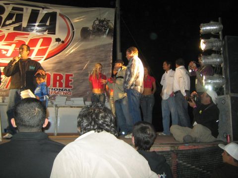1990 Ford Bronco - nighttime award presentation on a stage backed by a Baja 250 SCORE off-road racing banner, with a crowd watching.