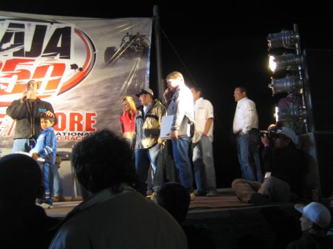 1990 Ford Bronco - People stand on a lit stage receiving awards in front of a Baja 250 SCORE racing banner as a crowd watches.