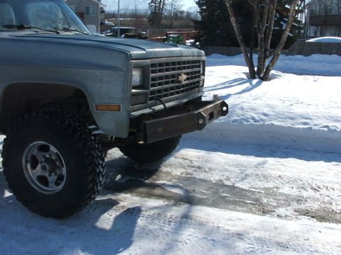 1990 Chevrolet Suburban - The front grille and headlights of a classic Chevrolet pickup truck are visible, with a custom bumper and snow on the ground.