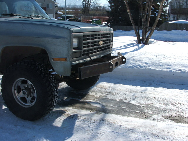Front of a light blue Chevrolet pickup truck with a custom metal bumper and large tires, parked in the snow