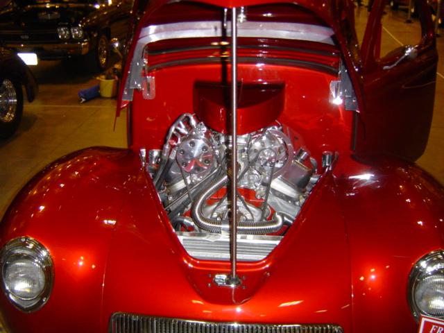 Custom red hot rod with engine exposed, hood open. Another car visible in background