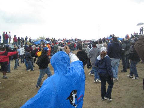 1990 Ford Bronco - person in a blue orca-printed rain poncho raises a camera to photograph a large crowd on a dusty open field.