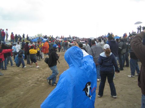 1990 Ford Bronco - Spectators in jackets, including one in a blue orca-print poncho, stand on a dusty field under a gray sky.