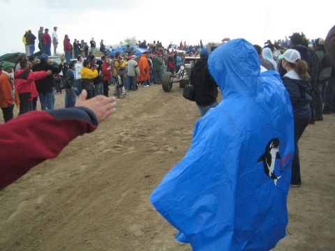 1990 Ford Bronco - spectator in a blue hooded poncho with an orca graphic watches an off-road race buggy pass a crowd on a dirt course under overcast skies.