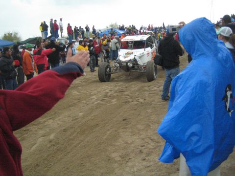 1990 Ford Bronco - white single-seat off-road race buggy drives along a dirt track past a crowd of bundled-up spectators on a gray day.