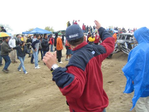 1990 Ford Bronco - man in a Castrol cap and red-and-navy jacket gestures with raised arms as spectators gather on a dirt field at an off-road race.