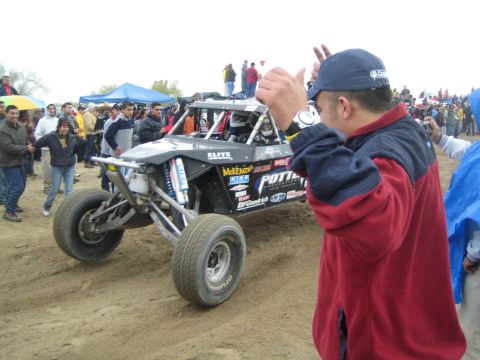 1990 Ford Bronco - black off-road race buggy with Potti, McKenzie and BFGoodrich sponsor decals sits on a dirt course as a man in a cap watches nearby.