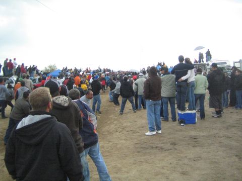 1990 Ford Bronco - large crowd of spectators in jackets and hoodies stands on a dirt field watching an event under heavy overcast skies.