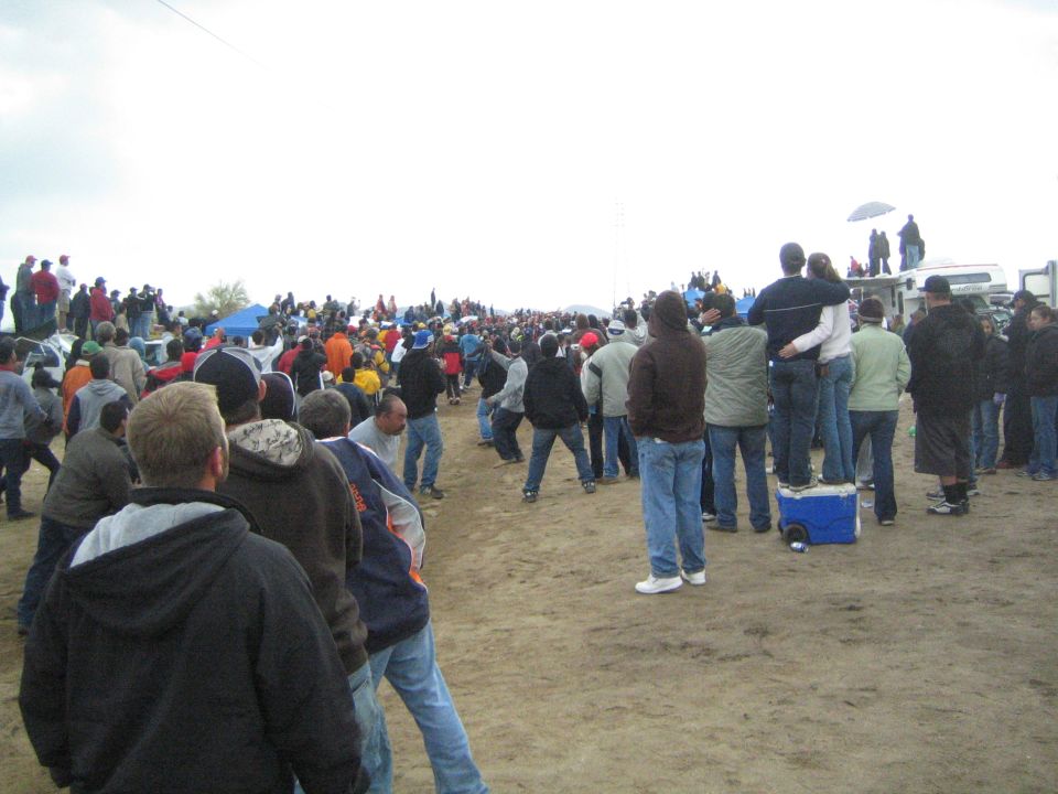Crowd of spectators in jackets standing on dirt field on overcast day