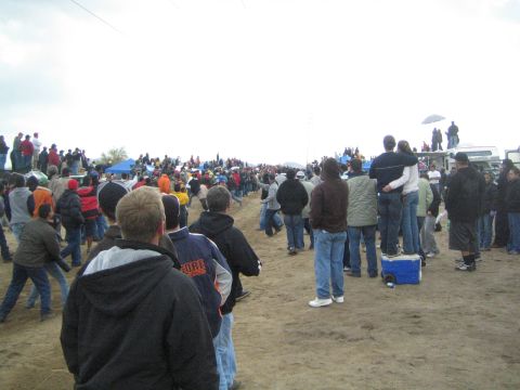 1990 Ford Bronco - Spectators in cold-weather clothing watch from a dirt field, with a couple standing on a cooler for a better view on a gray day.