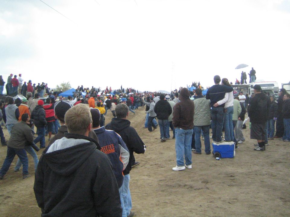 Spectators watching from a dirt field, couple standing on a cooler