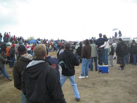 1990 Ford Bronco - crowd of spectators in dark jackets gathers on a dirt field watching an event, with a couple perched on a cooler under gray skies.