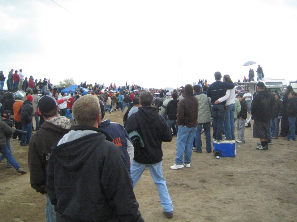 Crowd of bundled spectators on dirt field under gray sky
