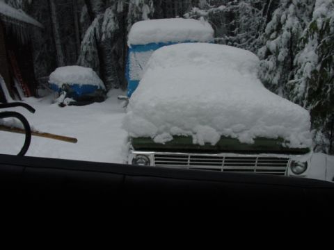 1986 Ford Bronco - A green vehicle, likely a truck or SUV, is buried in snow with only its front grille visible. Other snow-covered items are in the backgroun…