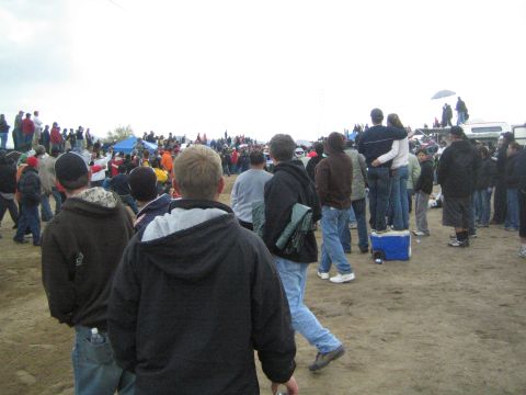1990 Ford Bronco - Spectators in jackets walk across a dirt field carrying gear and a cooler as they head toward a larger crowd on a gray day.