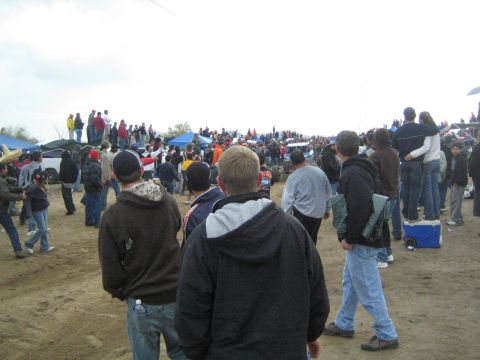 1990 Ford Bronco - crowd of spectators stands on a dirt field dotted with canopy tents, watching an event under a flat gray sky.