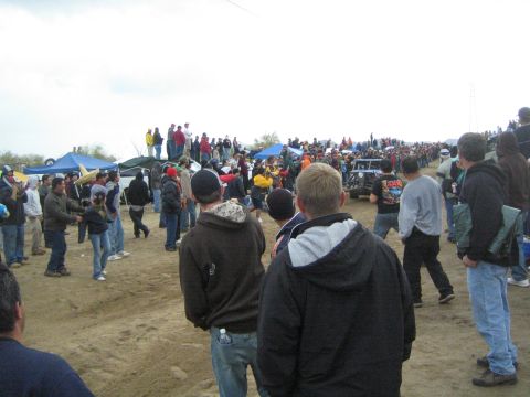1990 Ford Bronco - Spectators line a dirt course as a distant race vehicle approaches, with canopy tents and a large crowd in the background.