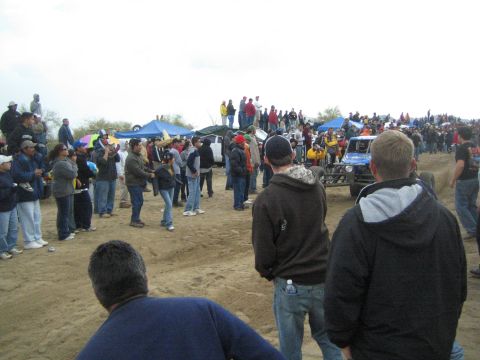 1990 Ford Bronco - crowd of spectators watches a blue and yellow off-road vehicle on a dirt course lined with canopy tents on an overcast day.