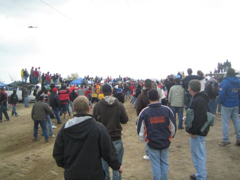 1990 Ford Bronco - helicopter hovers above a dirt field crowded with spectators in jackets watching an off-road race on an overcast day.