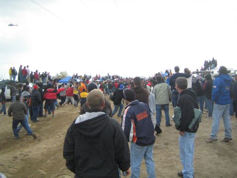 1990 Ford Bronco - helicopter flies over a large crowd of bundled spectators gathered on a dirt field at an off-road race under gray skies.