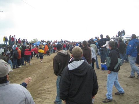 1990 Ford Bronco - Spectators in winter jackets stand on a dirt field with canopy tents and parked vehicles, watching an event on an overcast day.