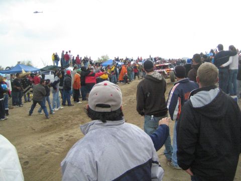 1990 Ford Bronco - crowd watches along a dirt course as a helicopter hovers and race vehicles gather in the distance on a gray day.