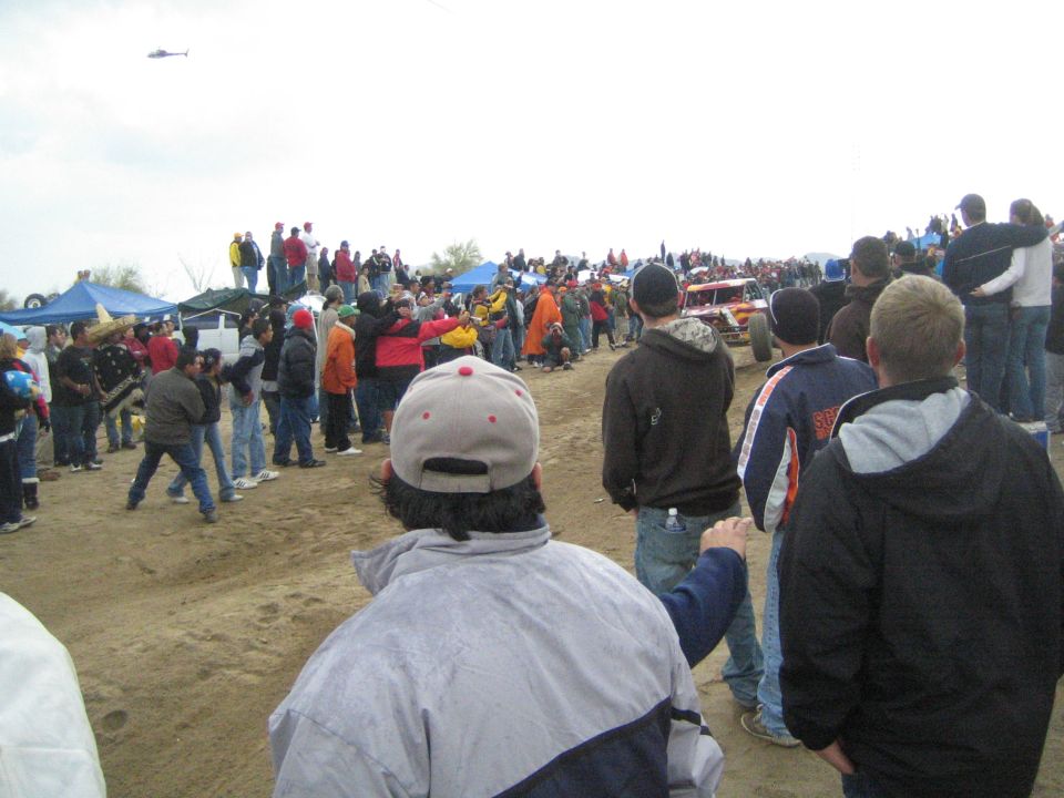 Crowd along dirt course with helicopter and race vehicles in distance