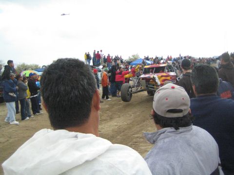1990 Ford Bronco - red off-road race buggy with flame paint drives along a dirt course surrounded by spectators as a helicopter flies overhead.