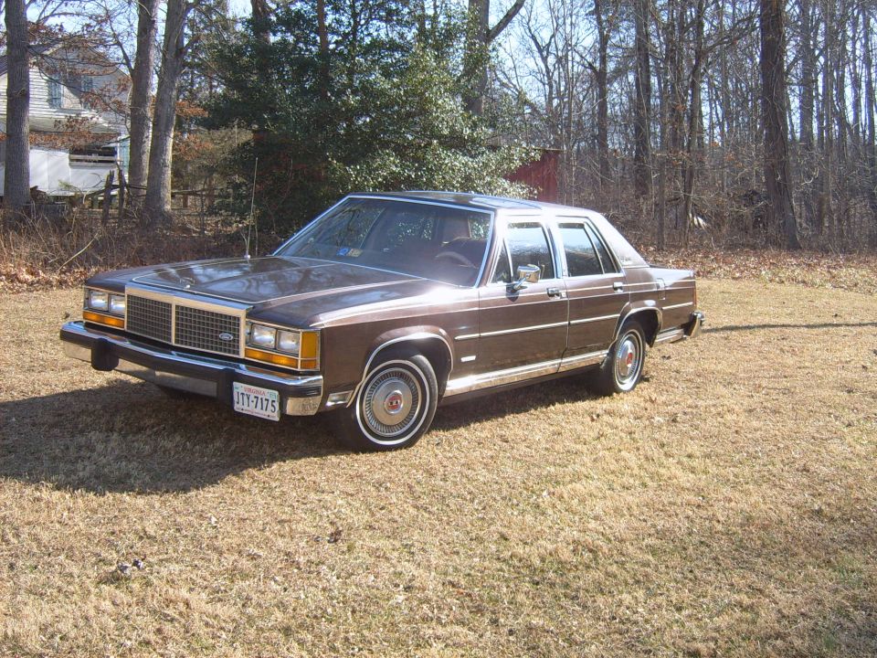 Brown 1980s Ford sedan with chrome trim, parked on dry grass