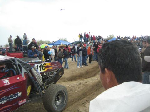 1990 Ford Bronco - red off-road race buggy numbered 116 with flame graphics rounds a dirt course as spectators line the track and a helicopter flies above.