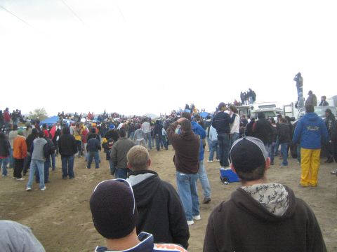 1990 Ford Bronco - large crowd of spectators in jackets and beanies fills a dirt field beside a parked RV, watching an event under overcast skies.