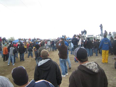 1990 Ford Bronco - dense crowd of spectators gathers on a dirt field while a person photographs from atop a parked RV on a gray day.