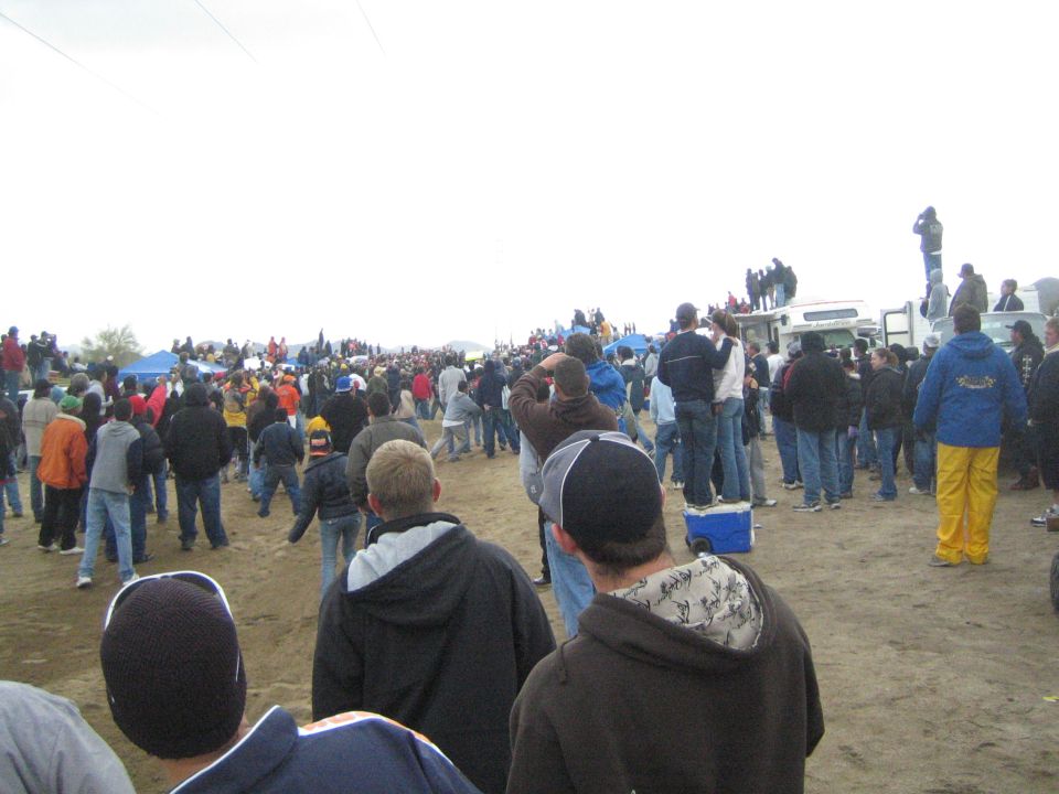Spectators spread across dirt field watching an event on overcast day
