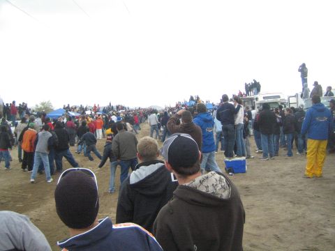 1990 Ford Bronco - crowd of spectators lines a dirt course beside a parked RV and canopy tents, watching an off-road event under overcast skies.