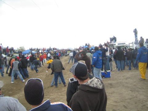 1990 Ford Bronco - large crowd of spectators in jackets and beanies stands on a dirt field watching an off-road race beside an RV under gray skies.