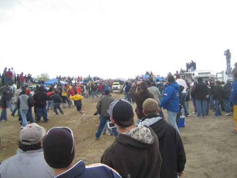 1990 Ford Bronco - Bundled-up spectators crowd a dirt off-road racecourse under an overcast sky, with blue canopy tents and trucks in the distance.