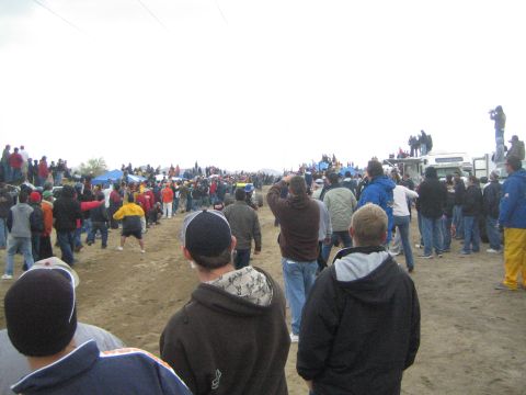 1990 Ford Bronco - dense crowd of spectators in cold-weather clothing lines a dirt road, watching an off-road race beneath a gray sky.