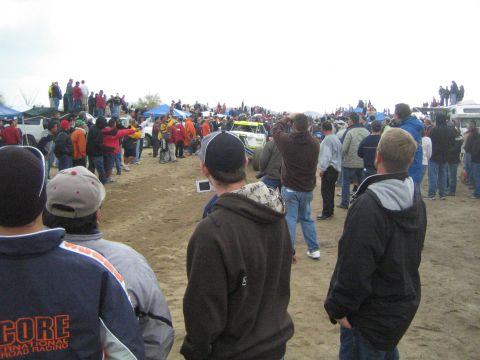 1990 Ford Bronco - small open-wheel off-road race car sits on a dirt course as spectators in jackets and caps watch under an overcast sky.