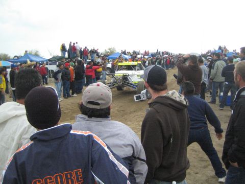 1990 Ford Bronco - yellow single-seat off-road buggy drives through a dirt course flanked by a crowd of spectators at a desert race.