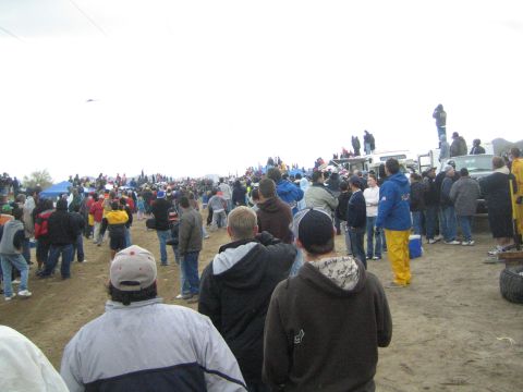 1990 Ford Bronco - broad crowd of spectators in jackets and caps gathers along a dirt off-road course beneath a hazy gray sky.
